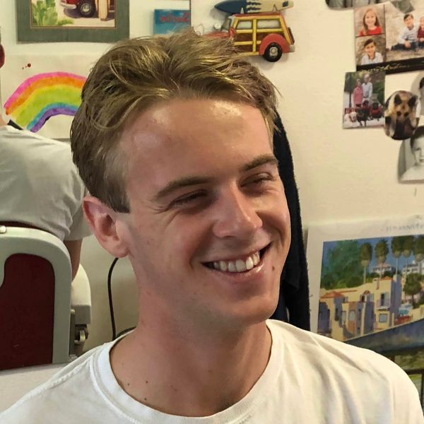 A young man with blonde hair smiling warmly indoors.