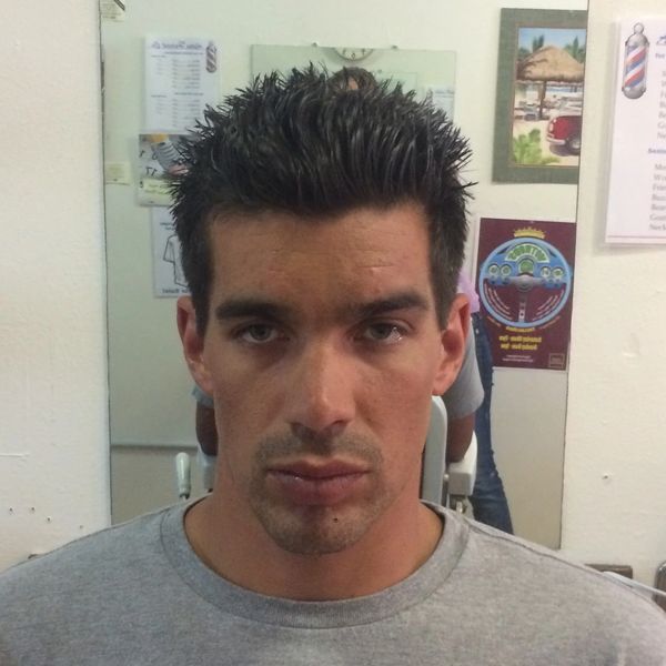 Man with spiked hair sitting in a barber shop with a clock behind him.