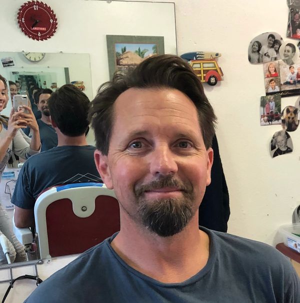 Man with a goatee smiling in a barber shop with a mirror reflecting a woman taking a photo.