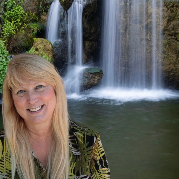 Smiling woman with blonde hair in front of a serene waterfall.