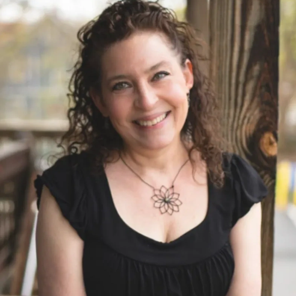 Smiling woman with curly hair in a black dress standing by a wooden post.