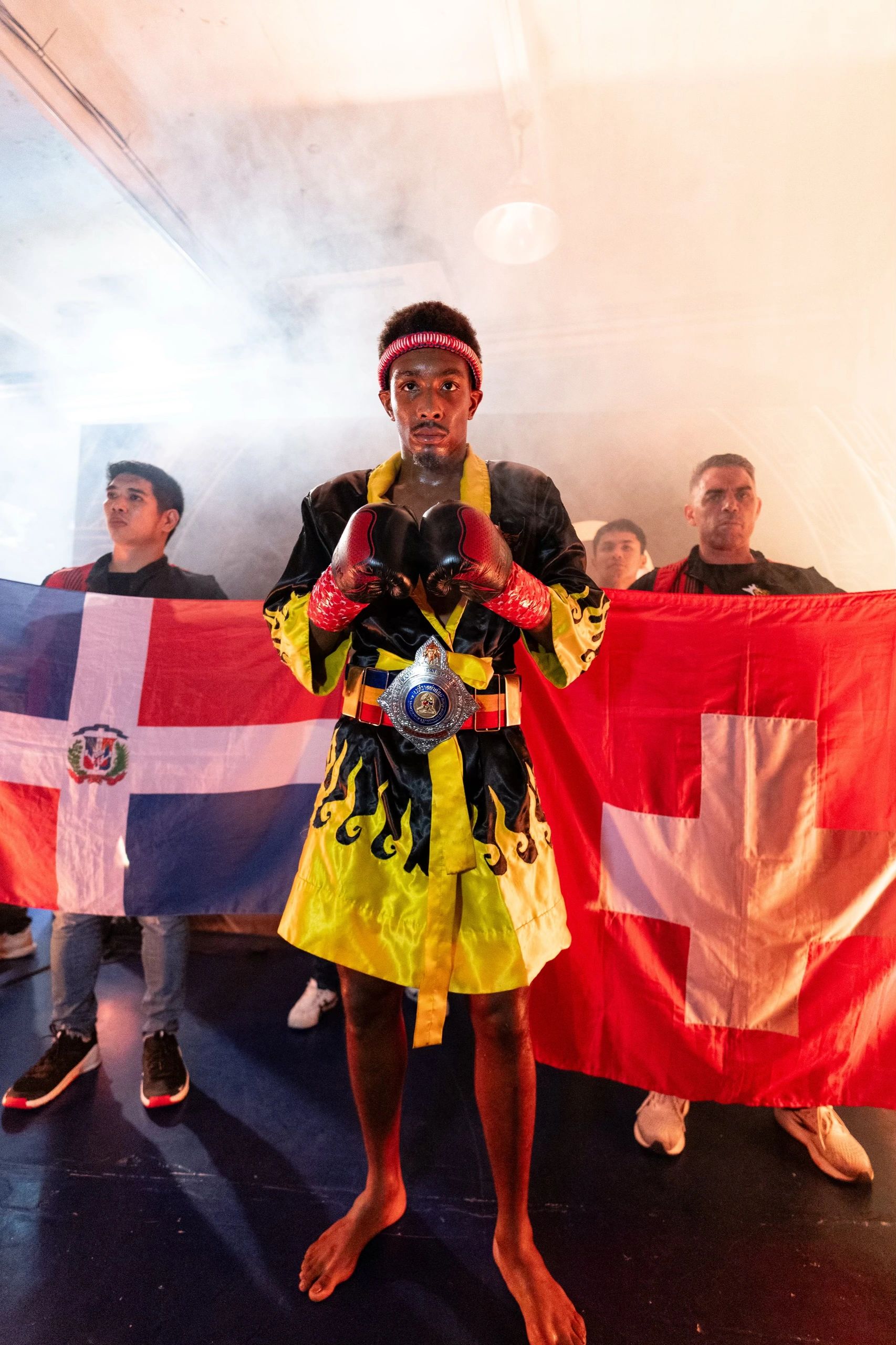 A focused boxer in yellow and black robe with red gloves poses confidently, flanked by supporters holding flags.