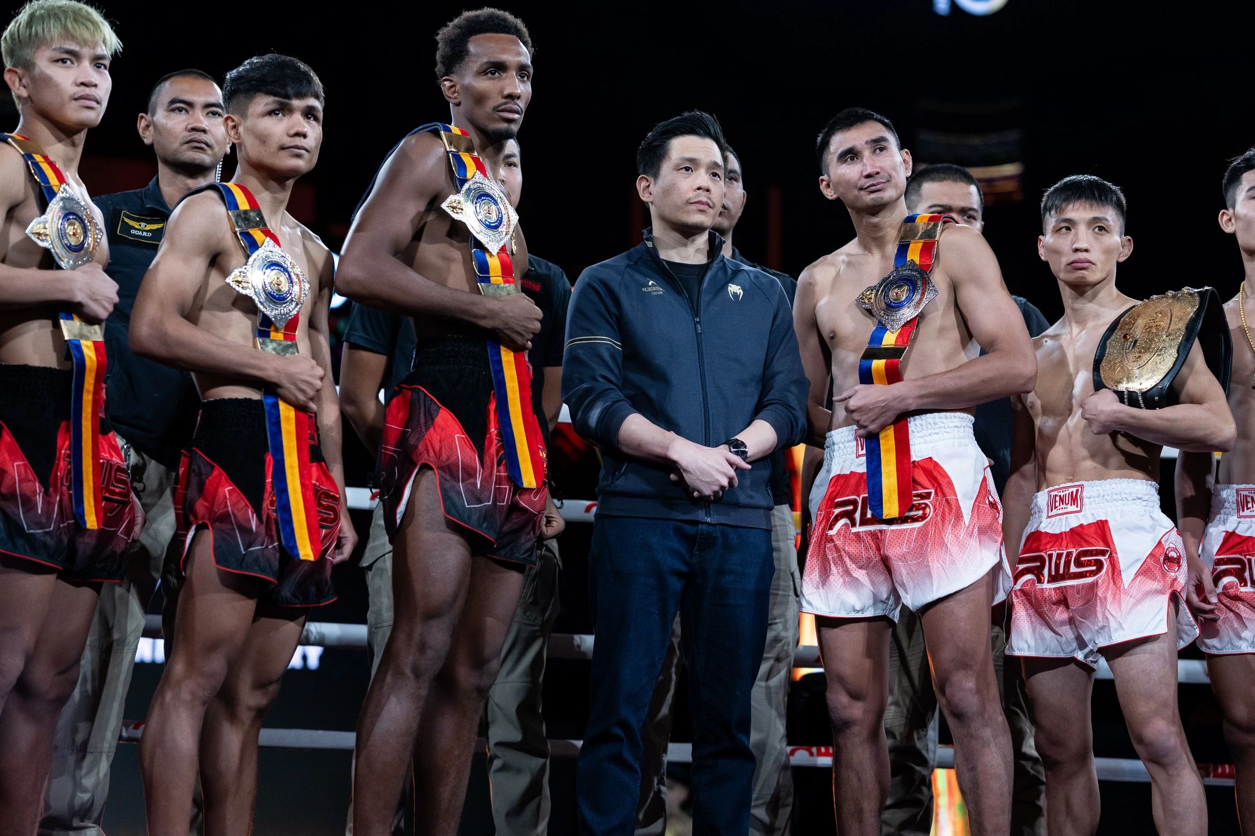 Group of Muay Thai fighters with championship belts standing together in a ring.