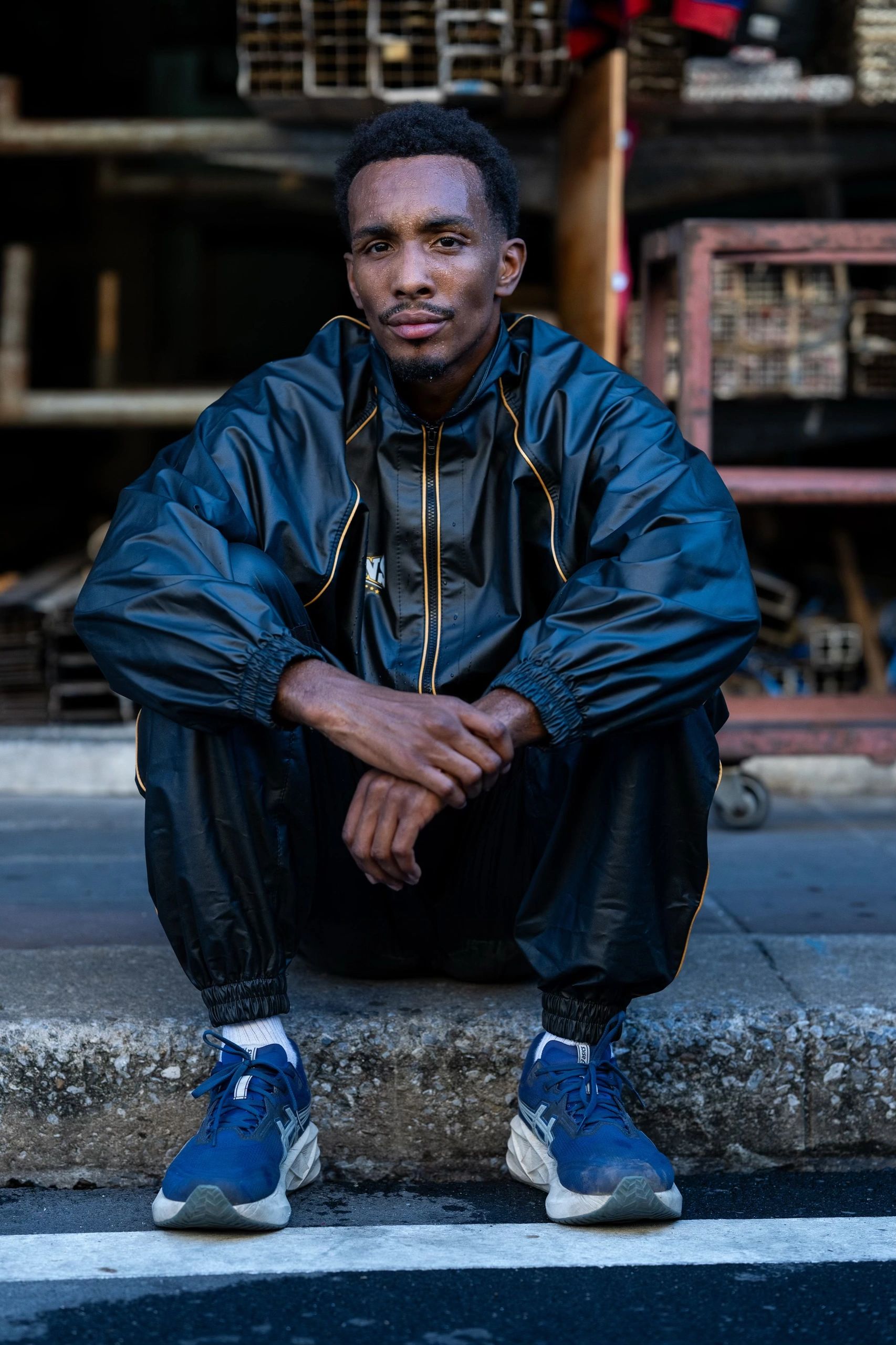 Man in black tracksuit and blue sneakers sitting on a curb.