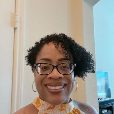 A smiling woman with glasses and curly hair wearing a yellow floral top.