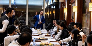 A man in a blue suit addressing a group at a formal dinner meeting.