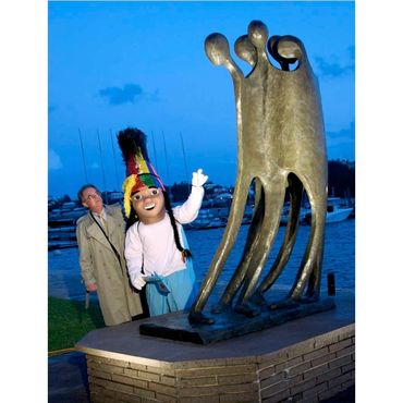 Person and mascot beside abstract sculpture by water.