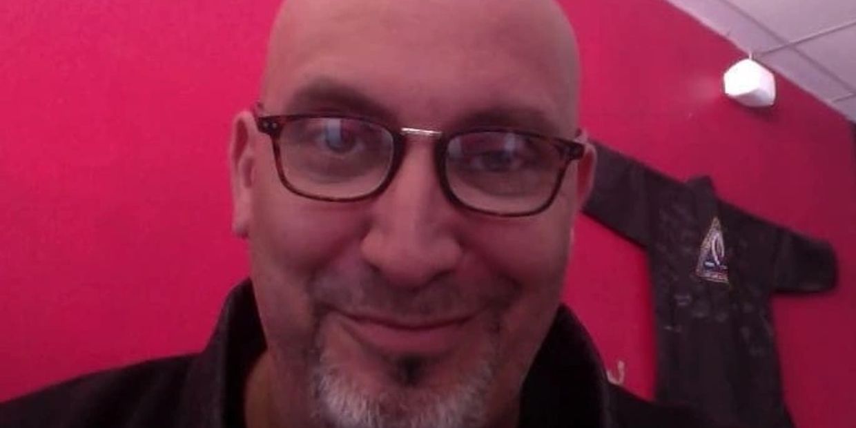 Smiling with glasses and goatee in front of a red wall.