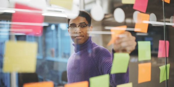 Man in glasses arranging colorful sticky notes on glass wall.