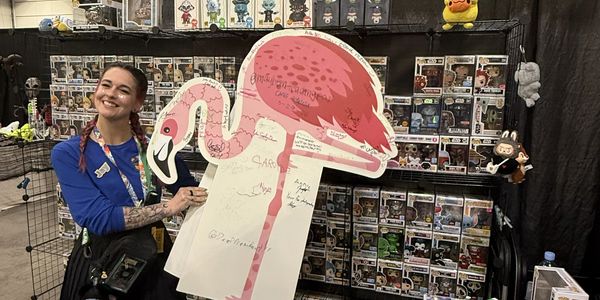 Smiling woman holding a large flamingo cutout at a collectible toy booth.