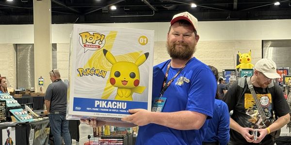 Man smiles holding a large Pikachu Funko Pop box at a convention.