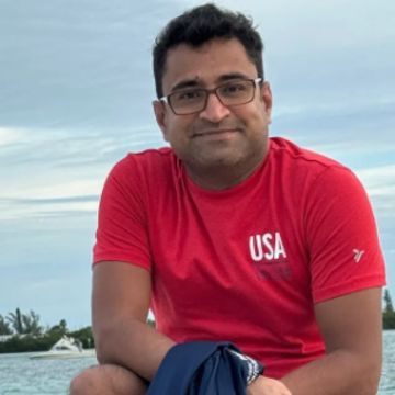 Man wearing a red USA t-shirt smiling outdoors near water.