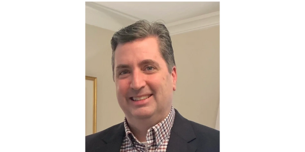 Man smiling, wearing a checkered shirt and dark blazer indoors.