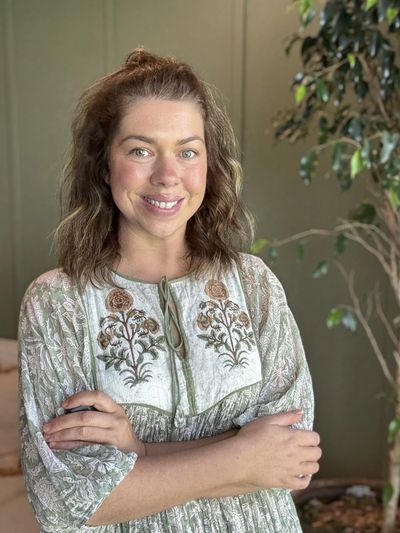 Smiling woman with wavy hair in a floral dress stands indoors with arms crossed.