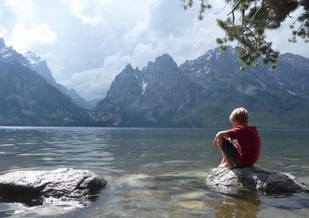 Child sitting on a rock by a lake with mountain view.