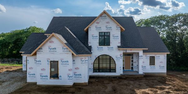 A newly constructed house wrapped in Tyvek weather barrier with black roofing under a blue sky.