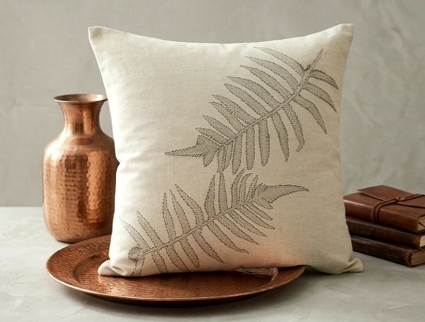 Decorative beige pillow with leaf design, placed on a copper plate with copper vase and stacked books.
