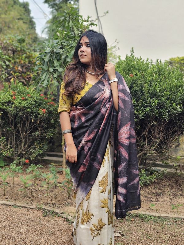 Woman in a floral saree standing outdoors with greenery in the background.