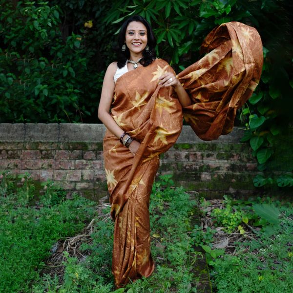Woman in a flowing brown saree stands amidst greenery with a smile.