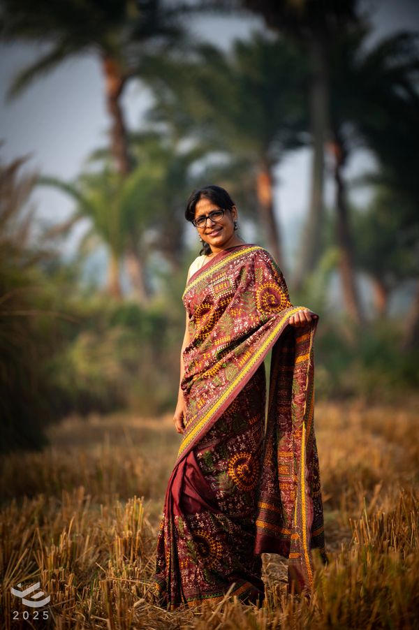 Woman in a traditional maroon sari stands in a field with a smile.