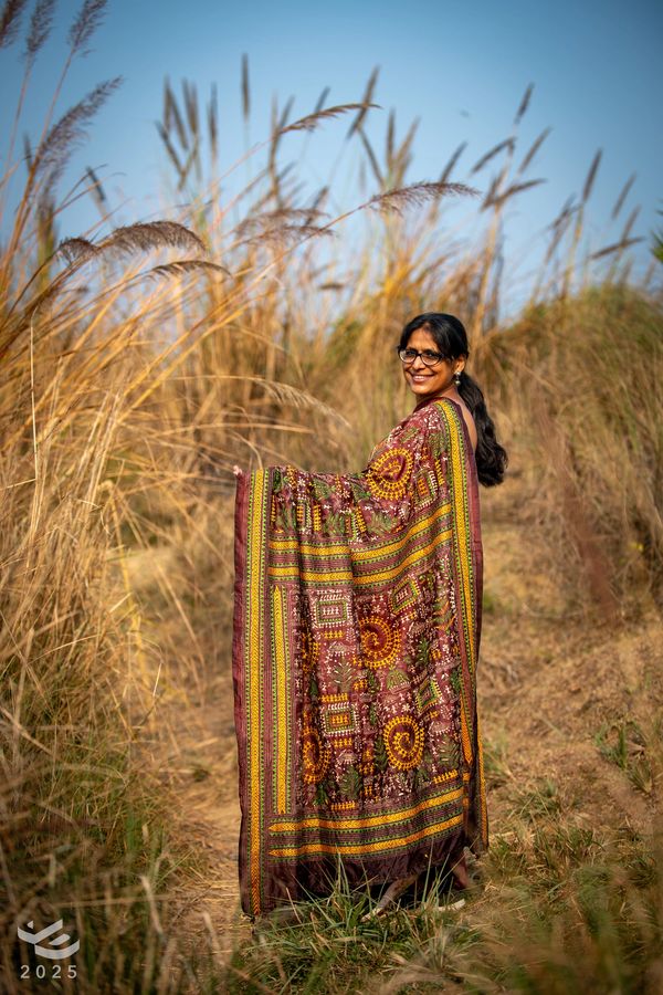 A woman in a colorful saree stands smiling in a grassy field under clear blue sky.