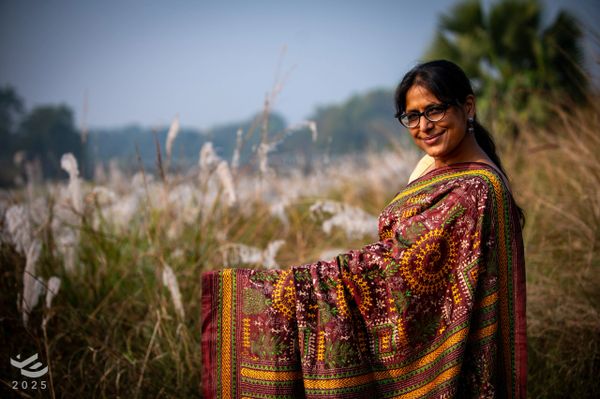 A woman in glasses smiling in a field, draped in a colorful traditional shawl.