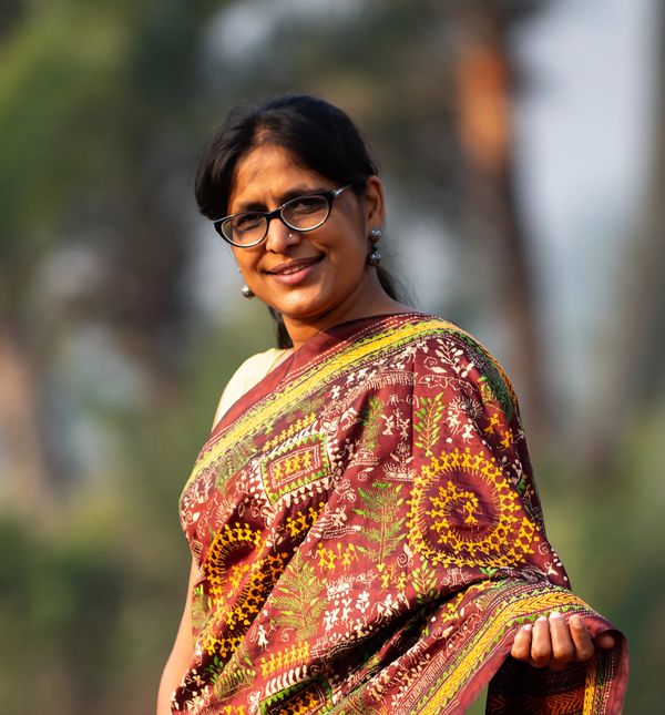 A woman smiling, wearing glasses and a colorful traditional saree outdoors.