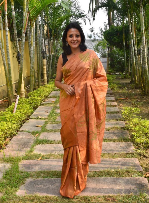 A woman in an orange saree stands on a stone path lined with greenery and palm trees.