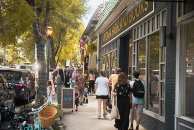 People walk along a lively urban street lined with shops and trees.
