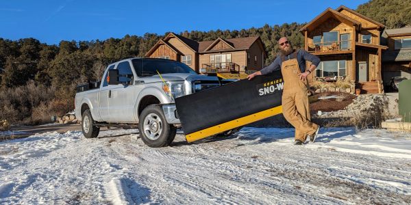 Man with snow plow attachment in front of a truck on a snowy driveway.
