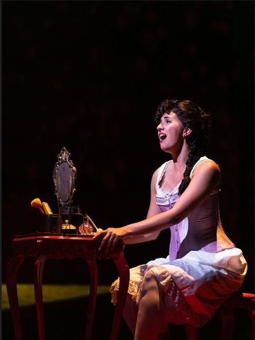 A woman in vintage attire sings passionately at a vanity table on stage.