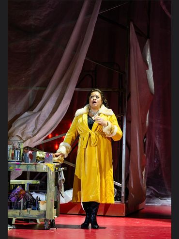 A woman in a yellow coat stands on a theatrical stage with dramatic curtains.