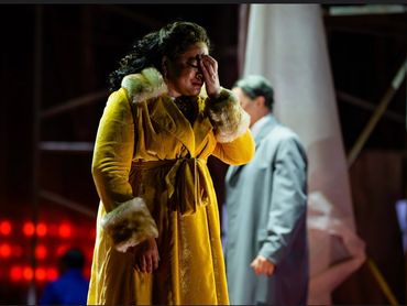 A woman in a yellow coat appears distressed on stage with a man in the background.