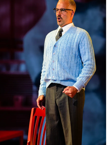 Man in blue cardigan and glasses performing on stage.