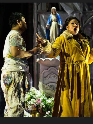 Two actors passionately performing on stage with a statue and flowers in the background.