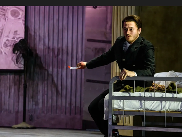A man in a dark coat holding a bloody knife, sitting on a bed in a dimly lit room.