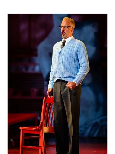 Man in blue cardigan and glasses performing on stage beside a red chair.