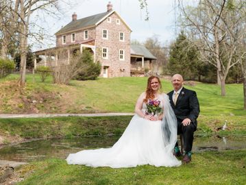 Newlywed couple posing outdoors near a pond with a stone house in the background.