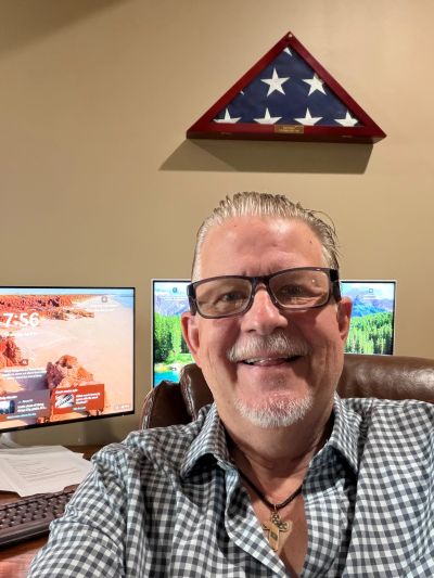 Man smiling in front of dual monitors with a folded American flag on the wall.