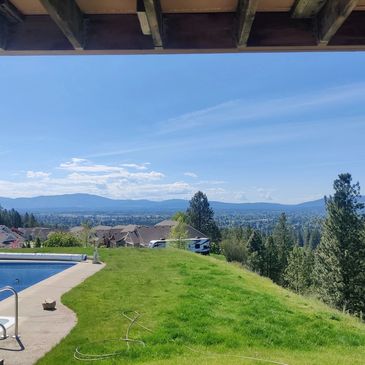 Scenic backyard with a pool, grassy hill, houses, and distant mountains under a clear blue sky.