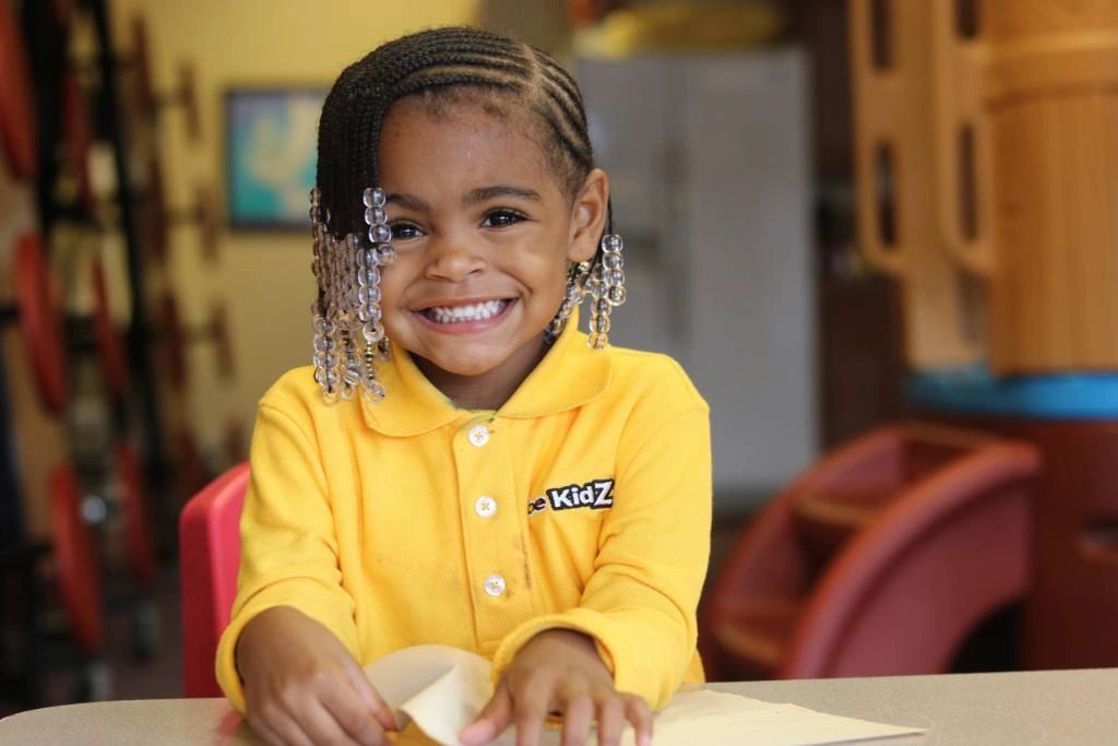 Smiling young girl with beaded braids wearing a yellow KidZ shirt.