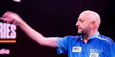 A man in a blue shirt throws a dart in a dimly lit setting.