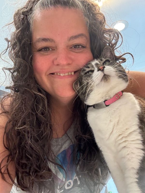 A woman with curly hair smiles while holding a white and gray cat wearing a pink collar.
