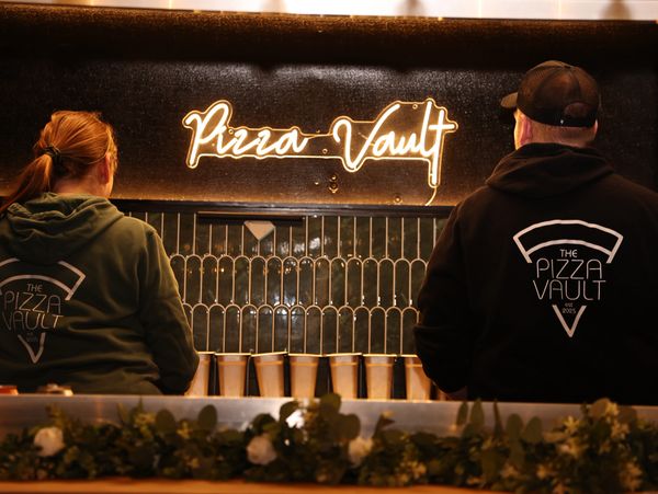 Two people in "The Pizza Vault" hoodies stand under a neon Pizza Vault sign.