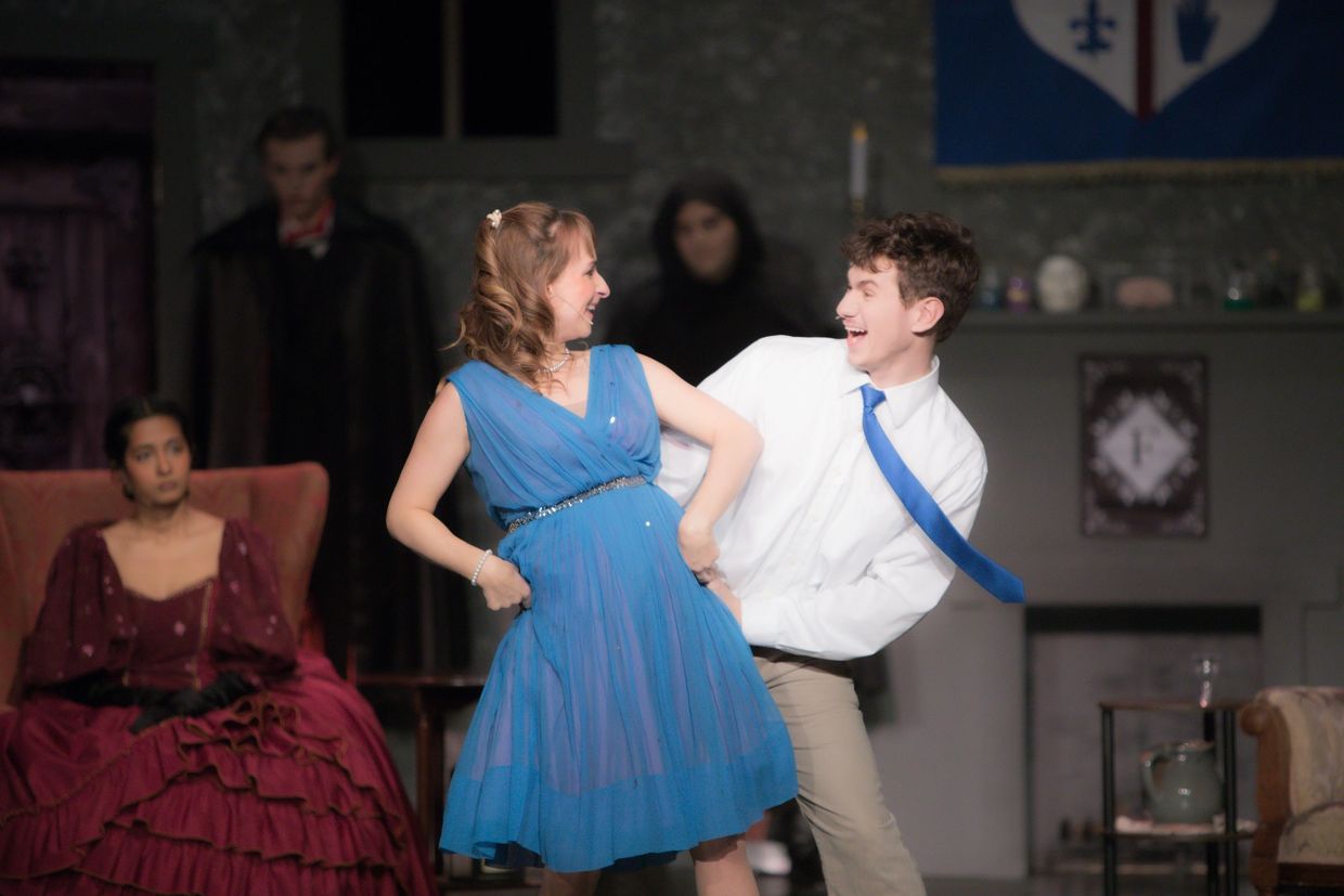 Two young actors joyfully dancing on stage with serious characters watching in the background.