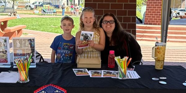 Two kids and a woman at a Dixon Community Theater booth outdoors.