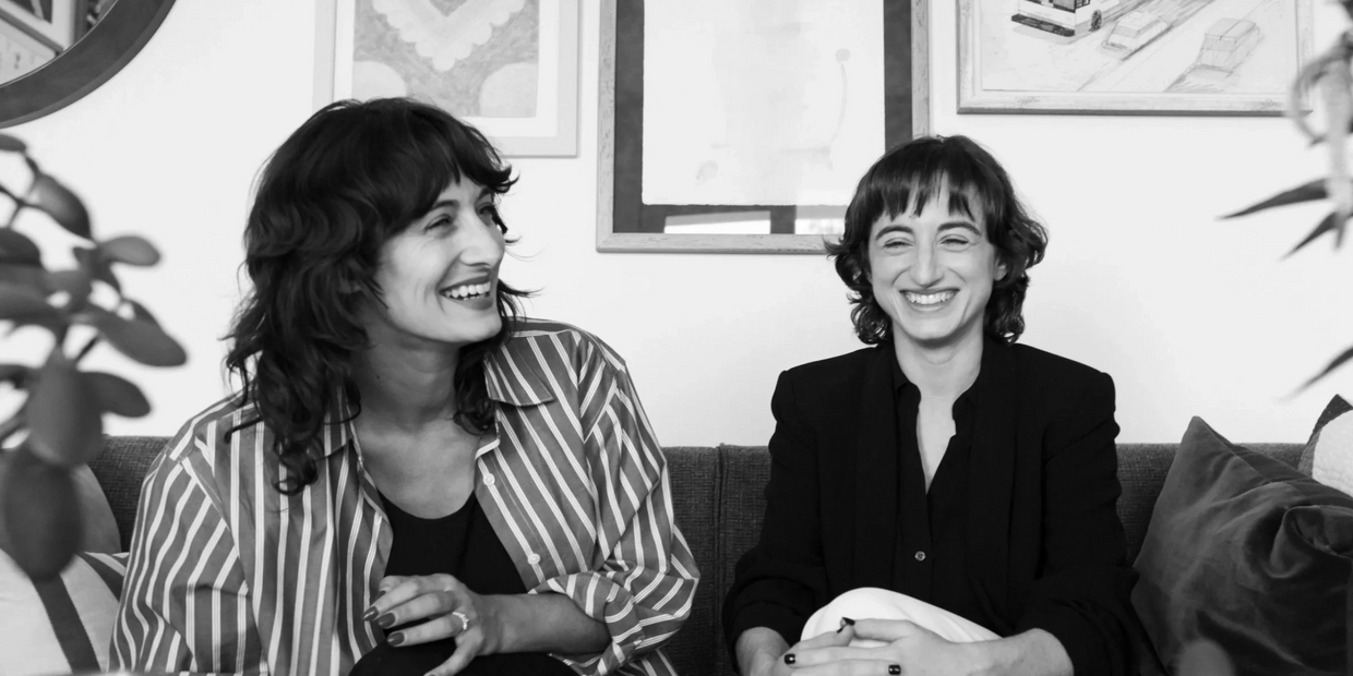 Two women laughing together on a couch in a cozy, art-filled room.