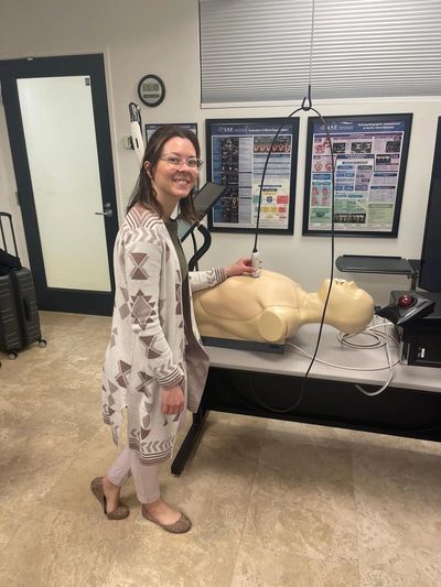 A woman smiling next to a CPR training mannequin in a classroom.