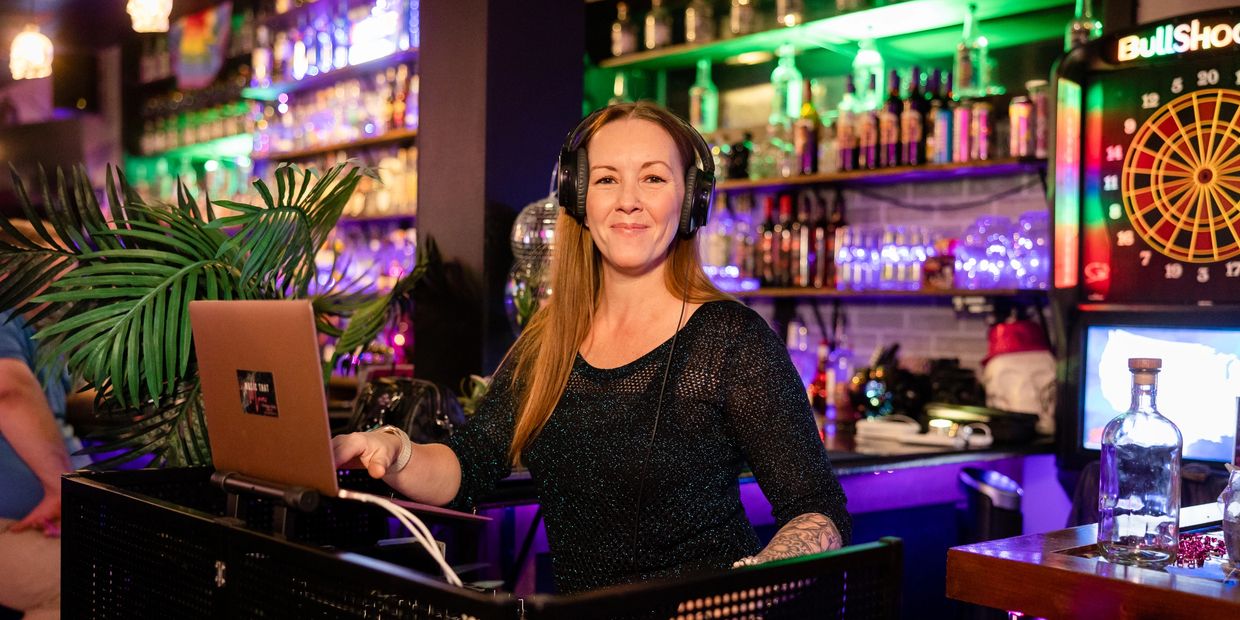 Smiling female DJ wearing headphones at a bar with colorful lights.