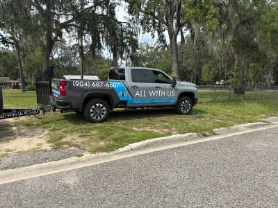 Pickup truck with "All With Us" business signage parked by a fence.